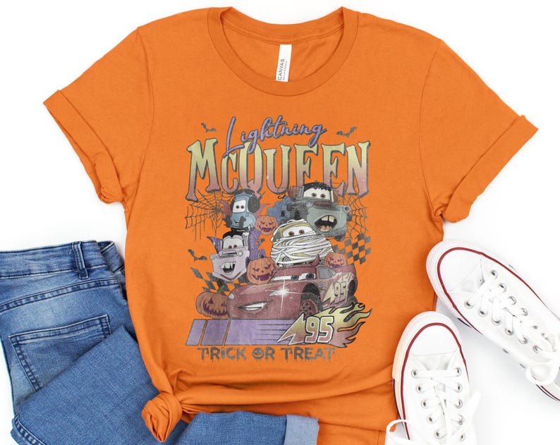May include: Orange t-shirt featuring a graphic of Lightning McQueen and other Cars characters in Halloween costumes. The text "Lightning McQueen" and "Trick or Treat" are also displayed. The shirt is paired with blue jeans and white sneakers.
