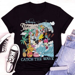 May include: Black t-shirt with a colorful graphic of Mickey Mouse and friends at Disney's Typhoon Lagoon water park. The graphic includes a wave, palm trees, a pineapple, and a beach ball. The text "Catch the Wave" is printed below the graphic.