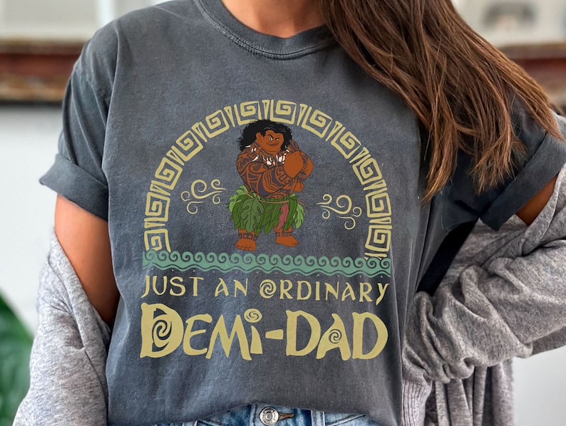 May include: Dark grey t-shirt with a cartoon character in a grass skirt and tattoos. The text "JUST AN ORDINARY DEMI-DAD" is in gold. Decorative elements surround the character, creating a themed design.