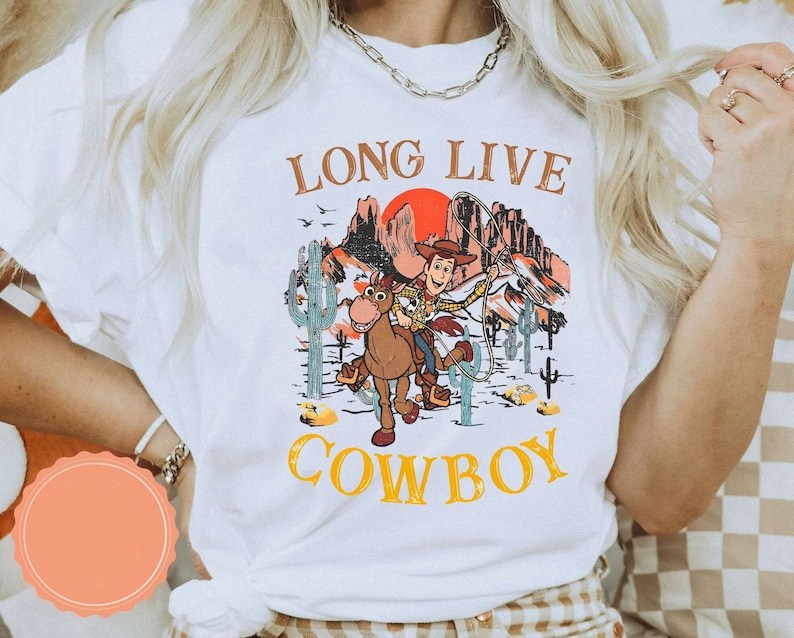 Pu&ograve; includere: Maglietta bianca con una grafica di Woody di Toy Story a cavallo in un paesaggio desertico. Il testo "Long Live Cowboy" &egrave; stampato sulla maglietta.