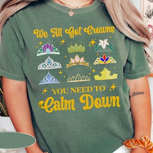 Op de afbeelding: Groen T-shirt met de tekst "We All Got Crowns You Need To Calm Down" in geel. Het shirt heeft een verscheidenheid aan kleurrijke kronen.