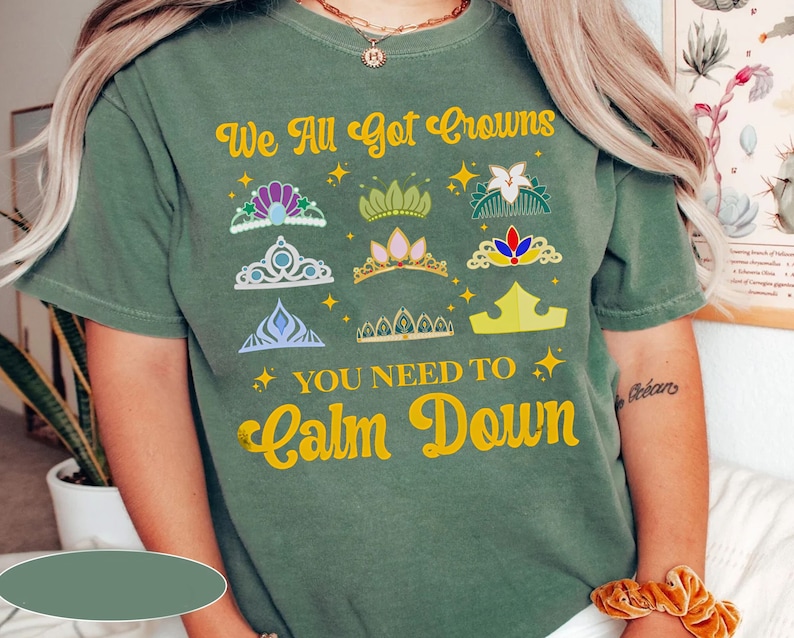 Op de afbeelding: Groen T-shirt met de tekst "We All Got Crowns You Need To Calm Down" in geel. Het shirt heeft een verscheidenheid aan kleurrijke kronen.