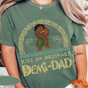 May include: A green t-shirt featuring a cartoon character and the text "JUST AN ORDINARY DEMI-DAD" in yellow. The character is brown with tattoos and wearing a leaf skirt. The shirt has a crew neck and short sleeves.