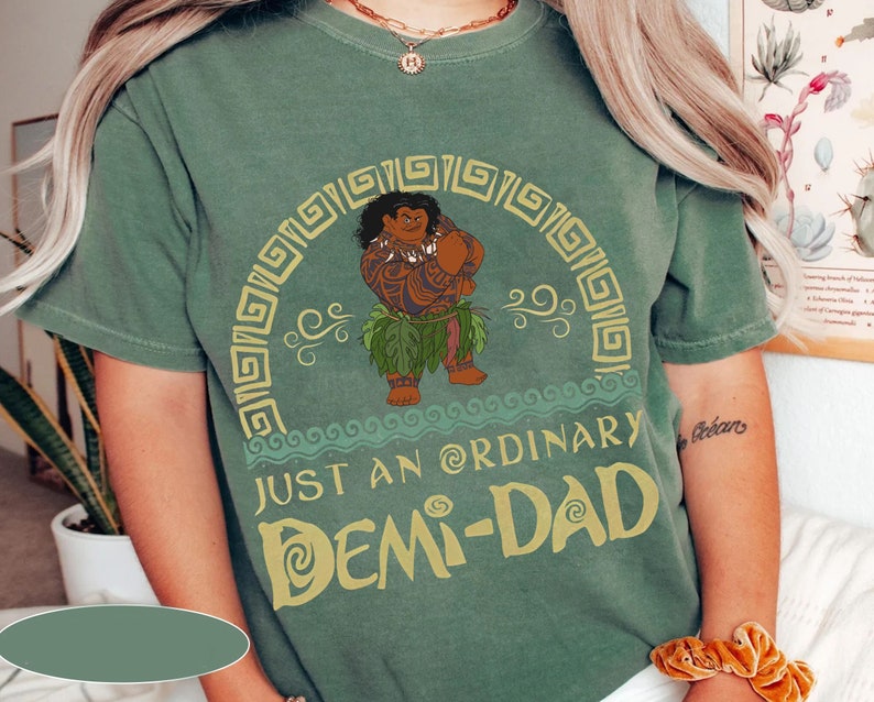 May include: A green t-shirt featuring a cartoon character and the text "JUST AN ORDINARY DEMI-DAD" in yellow. The character is brown with tattoos and wearing a leaf skirt. The shirt has a crew neck and short sleeves.