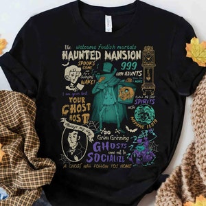 May include: Black t-shirt with a colorful graphic design featuring a haunted mansion theme. The design includes a ghost, a clock, and the text "The Welcome Foolish Mortals Haunted Mansion 999 Happy Haunts".