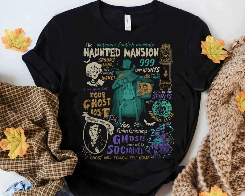 May include: Black t-shirt with a colorful graphic design featuring a haunted mansion theme. The design includes a ghost, a clock, and the text "The Welcome Foolish Mortals Haunted Mansion 999 Happy Haunts".