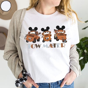May include: White t-shirt with a graphic of three orange tow trucks with Mickey Mouse ears. The tow trucks are labeled "Tow Mater".