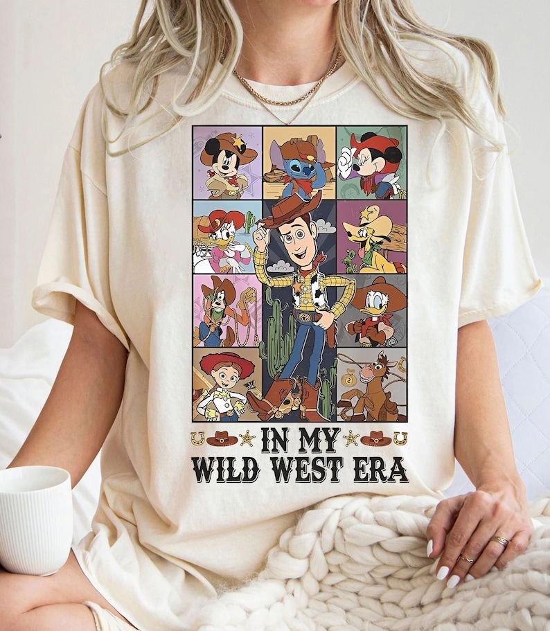 Puede incluir: Una camiseta beige con un gr&aacute;fico de dibujos animados de varios personajes de Disney en un entorno del oeste. El texto "In My Wild West Era" est&aacute; impreso debajo del gr&aacute;fico.