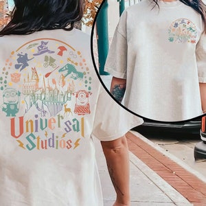 Könnte beinhalten: Ein weißes T-Shirt mit einer bunten Grafik beliebter Universal Studios-Attraktionen, darunter Hogwarts, Minions und Jurassic Park. Der Text "Universal Studios" ist unter der Grafik aufgedruckt.