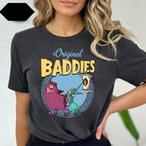 May include: A dark grey t-shirt featuring a graphic print of two cartoon characters. The characters are a purple creature with a crown and a green creature with a long nose. The text "Original Baddies" is printed in yellow above the characters.