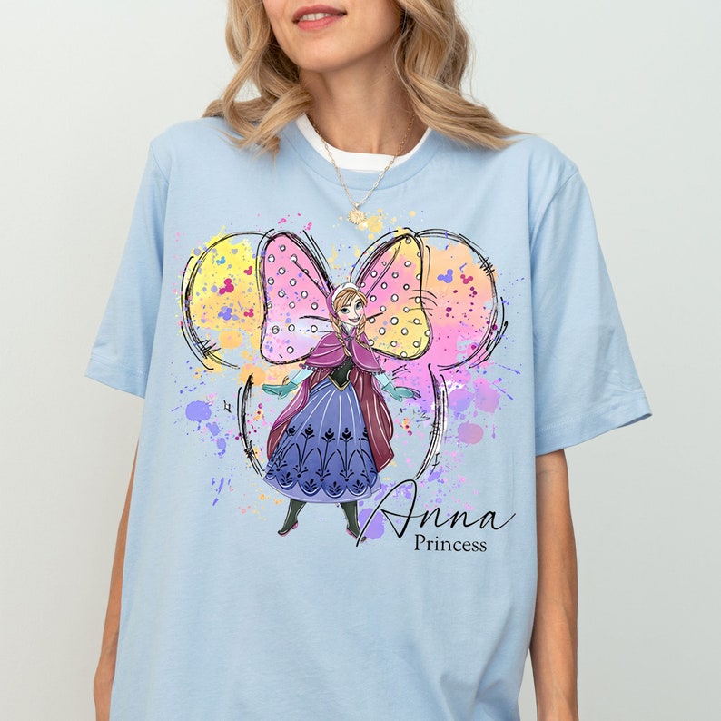 Peut inclure: T-shirt bleu clair avec un motif color&eacute; d'Anna de La Reine des Neiges portant un n&oelig;ud rose et jaune. Le motif est entour&eacute; d'&eacute;claboussures de peinture color&eacute;es. Le texte "Anna Princess" est imprim&eacute; sous le motif.