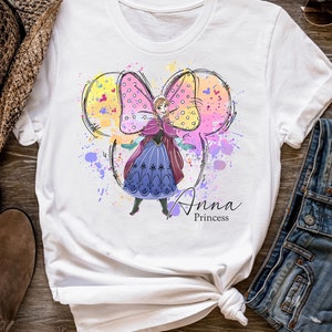 Peut inclure: T-shirt blanc avec une illustration aquarelle d'Anna de La Reine des Neiges portant une robe violette et un n&oelig;ud rose. Le n&oelig;ud a la forme des oreilles de Mickey Mouse. Le texte "Anna Princess" est imprim&eacute; sous l'illustration.