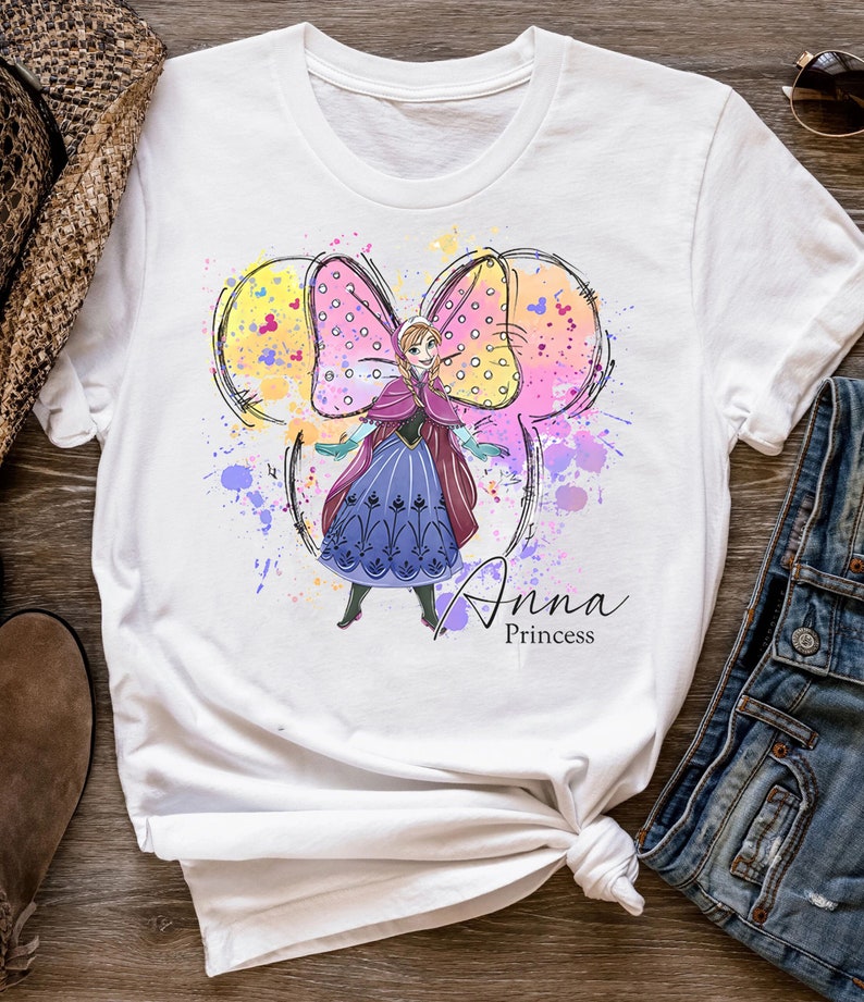 Peut inclure: T-shirt blanc avec une illustration aquarelle d'Anna de La Reine des Neiges portant une robe violette et un n&oelig;ud rose. Le n&oelig;ud a la forme des oreilles de Mickey Mouse. Le texte "Anna Princess" est imprim&eacute; sous l'illustration.