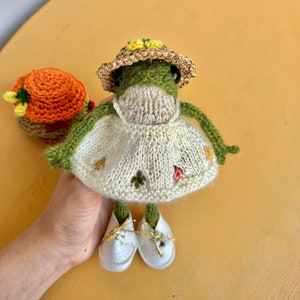 May include: A green crocheted frog wearing a straw hat with yellow flowers, a white dress with embroidered flowers, and white shoes with gold accents.