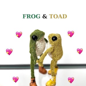 May include: Two knitted frog plushies, one green and one yellow, are holding hands and wearing shoes. The plushies are on a glass surface with pink hearts around them. The text "FROG & TOAD" is at the top of the image.