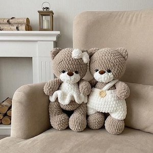 May include: Two handmade teddy bears sit on a beige armchair. The bears are light brown with white accents. One bear wears a white dress and bow, the other wears white overalls. The bears have brown eyes and noses.