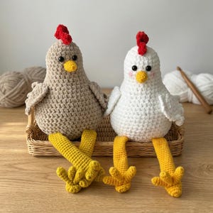 Könnte beinhalten: Zwei handgefertigte Häkelhühner. Eines ist beige, das andere ist weiß. Beide haben rote Kämme, gelbe Schnäbel und gelbe Füße. Sie sitzen in einem kleinen Weidenkorb auf einer Holzoberfläche. Die Hühner sind etwa 15 cm groß.