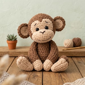 Puede incluir: Un mono de ganchillo hecho a mano en tonos marrón y beige. El peluche tiene una cara sonriente, orejas grandes y está sentado sobre una superficie de madera. Una pequeña maceta con suculentas y ovillos de lana están en el fondo.