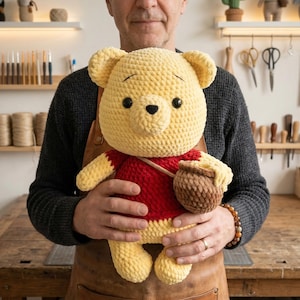 May include: A handmade, yellow crochet bear with a red shirt and a brown honey pot. The bear has black eyes and is being held by a person wearing a dark gray sweater and a brown apron. The bear is a plush toy.