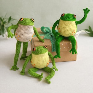 May include: Three handmade crochet frog toys in shades of green, yellow, and beige. The frogs have orange eyes and are posed in different positions, with one sitting on a gift box. The frogs have a whimsical, playful design.