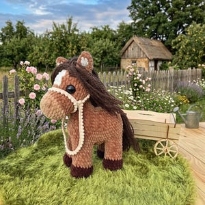 Puede incluir: Un caballo marrón de peluche hecho a mano, con una marca blanca y una melena y cola marrón oscuro. El caballo tiene un cabestro color crema y está sobre una alfombra verde y peluda. Un pequeño carro de madera está detrás del caballo, con un fondo rural.