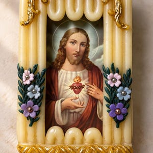 May include: A rectangular religious plaque featuring a central image of the Sacred Heart of Jesus. The frame is constructed from cream-coloured, ribbed wax-like material, decorated with gold filigree accents and small pink, blue, and purple flowers.