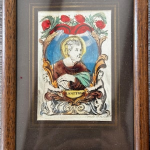 May include: A framed religious print featuring a central portrait of a bearded figure with a yellow halo, set against a blue oval background. The image includes red floral accents, two small cherub figures, and a banner labeled S. MATTHIAS.