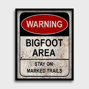 Puede incluir: Un letrero rectangular enmarcado en negro, con un diseño envejecido. La parte superior muestra un óvalo rojo con la palabra "WARNING" en blanco. Debajo, se lee "BIGFOOT AREA" y "STAY ON MARKED TRAILS", con un aspecto vintage.