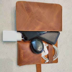 May include: A brown leather tech organizer, open to reveal a smartphone, charger, mouse, and cables. The organizer has a flap closure and a strap with a buckle. The leather has a textured appearance.