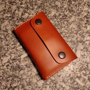 May include: A brown leather wallet with a snap closure. The rectangular wallet has white stitching along the edges. Two black snaps secure the flap. The leather has a smooth texture.
