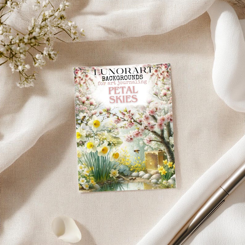 May include: A rectangular card with the text "LUNORART BACKGROUNDS for art journaling PETAL SKIES" features a floral illustration with daffodils, cherry blossoms, and a small bee. The card is on a cream-colored surface with a pen and a white fabric.