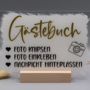 May include: A rectangular clear acrylic sign with a white painted brushstroke background, featuring the gold text G&auml;stebuch, a camera icon, and black text reading FOTO KNIPSEN, FOTO EINKLEBEN, and NACHRICHT HINTERLASSEN, supported by a light wood base.