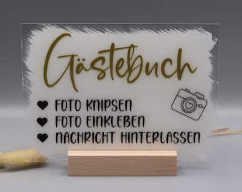 May include: A rectangular clear acrylic sign with a white painted brushstroke background, featuring the gold text Gästebuch, a camera icon, and black text reading FOTO KNIPSEN, FOTO EINKLEBEN, and NACHRICHT HINTERLASSEN, supported by a light wood base.