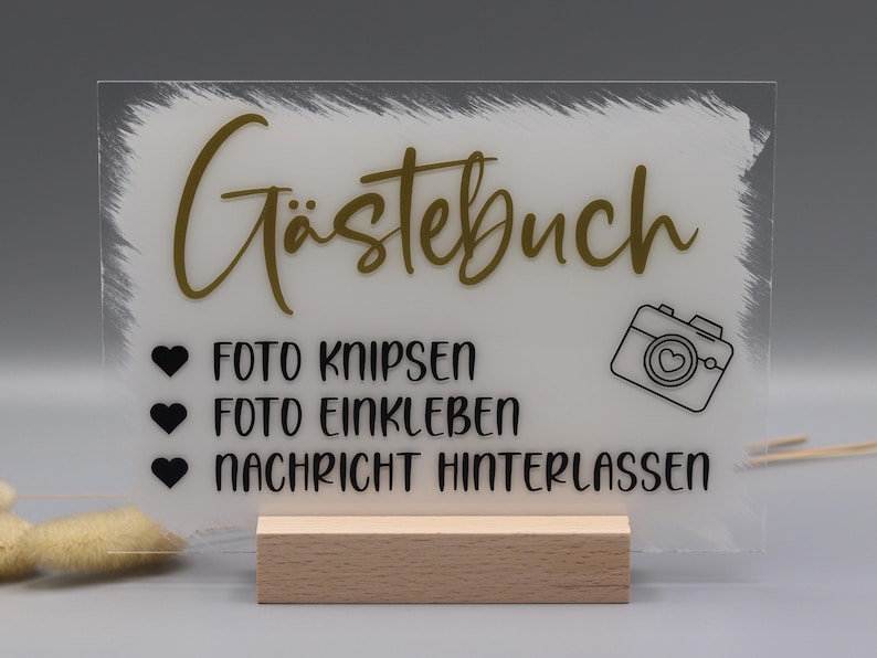 May include: A rectangular clear acrylic sign with a white painted brushstroke background, featuring the gold text G&auml;stebuch, a camera icon, and black text reading FOTO KNIPSEN, FOTO EINKLEBEN, and NACHRICHT HINTERLASSEN, supported by a light wood base.