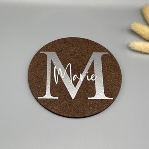 Puede incluir: Un posavasos redondo marr&oacute;n con una gran letra "M" plateada y el nombre "Marie" en escritura blanca. El posavasos est&aacute; hecho de un material similar al fieltro. Flores secas decorativas en el fondo.