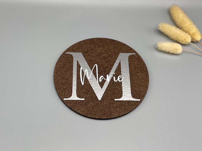 Puede incluir: Un posavasos redondo marr&oacute;n con una gran letra "M" plateada y el nombre "Marie" en escritura blanca. El posavasos est&aacute; hecho de un material similar al fieltro. Flores secas decorativas en el fondo.
