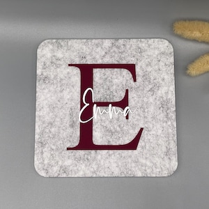 Puede incluir: Un posavasos cuadrado de fieltro gris con una gran letra "E" burdeos y el nombre "Emma" en escritura blanca. El posavasos est&aacute; sobre una superficie gris, con flores secas en la esquina superior derecha.