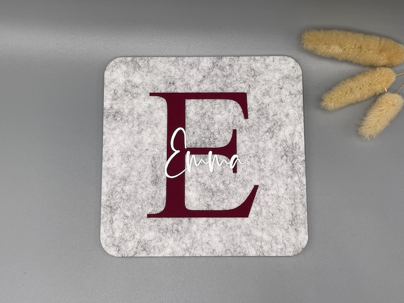 Puede incluir: Un posavasos cuadrado de fieltro gris con una gran letra "E" burdeos y el nombre "Emma" en escritura blanca. El posavasos est&aacute; sobre una superficie gris, con flores secas en la esquina superior derecha.