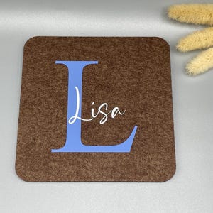 Puede incluir: Un posavasos cuadrado de fieltro marr&oacute;n con una gran letra "L" azul y el nombre "Lisa" en escritura blanca. El posavasos es de color marr&oacute;n liso con un aspecto texturizado. Flores secas en el fondo.