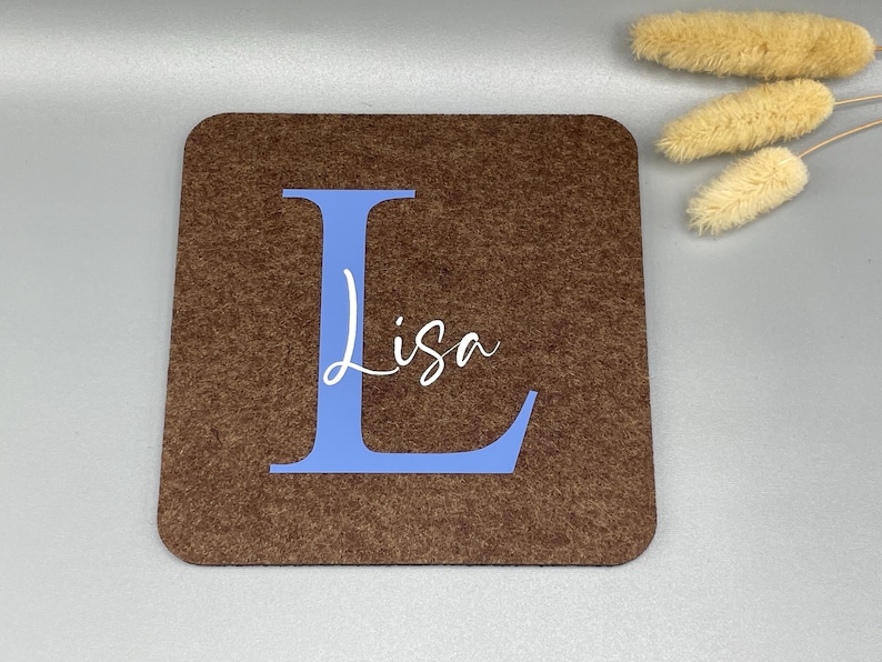 Puede incluir: Un posavasos cuadrado de fieltro marr&oacute;n con una gran letra "L" azul y el nombre "Lisa" en escritura blanca. El posavasos es de color marr&oacute;n liso con un aspecto texturizado. Flores secas en el fondo.