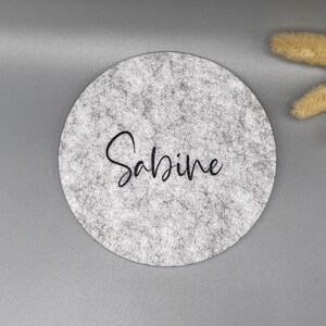 Puede incluir: Un posavasos redondo de fieltro gris con el nombre "Sabine" en escritura negra. El posavasos tiene una textura moteada y est&aacute; sobre un fondo gris. Trigo decorativo seco en la esquina superior derecha.