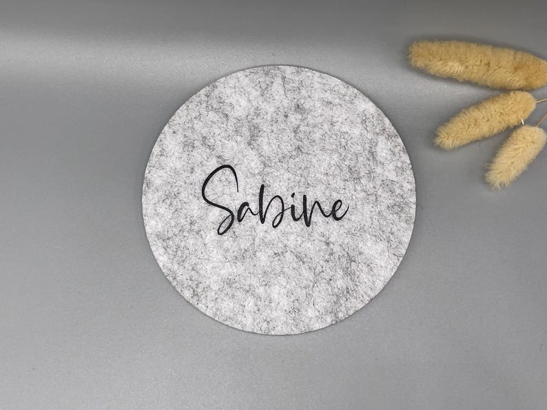 Puede incluir: Un posavasos redondo de fieltro gris con el nombre "Sabine" en escritura negra. El posavasos tiene una textura moteada y est&aacute; sobre un fondo gris. Trigo decorativo seco en la esquina superior derecha.