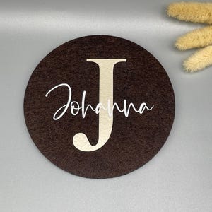 Puede incluir: Un posavasos redondo de fieltro marr&oacute;n oscuro con el nombre "Johanna" y la letra "J" en beige claro. El nombre est&aacute; escrito en cursiva. Flores secas en el fondo.