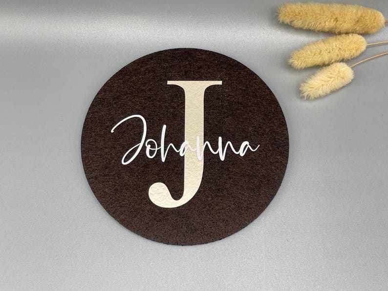 Puede incluir: Un posavasos redondo de fieltro marr&oacute;n oscuro con el nombre "Johanna" y la letra "J" en beige claro. El nombre est&aacute; escrito en cursiva. Flores secas en el fondo.