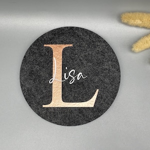 Puede incluir: Un posavasos redondo de fieltro gris oscuro con una gran letra "L" en oro rosa y el nombre "Lisa" en escritura blanca. Un art&iacute;culo decorativo para el hogar o la oficina.