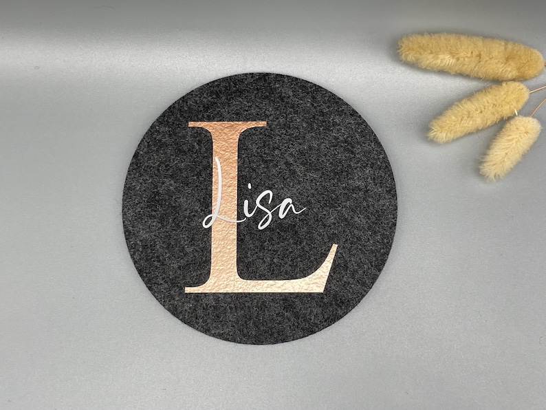 Puede incluir: Un posavasos redondo de fieltro gris oscuro con una gran letra "L" en oro rosa y el nombre "Lisa" en escritura blanca. Un art&iacute;culo decorativo para el hogar o la oficina.