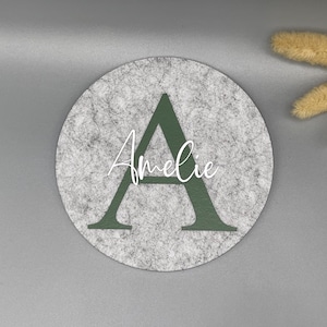 Puede incluir: Un posavasos redondo de fieltro gris con una gran letra "A" verde oscuro y el nombre "Amelie" en escritura blanca. El posavasos est&aacute; sobre una superficie gris, con flores secas en la esquina superior derecha.