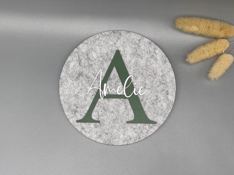 Puede incluir: Un posavasos redondo de fieltro gris con una gran letra "A" verde oscuro y el nombre "Amelie" en escritura blanca. El posavasos est&aacute; sobre una superficie gris, con flores secas en la esquina superior derecha.