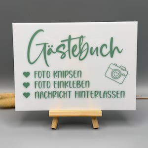 May include: A rectangular white acrylic sign featuring sage green text that reads G&auml;stebuch, Foto knipsen, Foto einkleben, and Nachricht hinterlassen. The sign includes three green heart icons and a camera graphic, displayed on a small wooden easel.