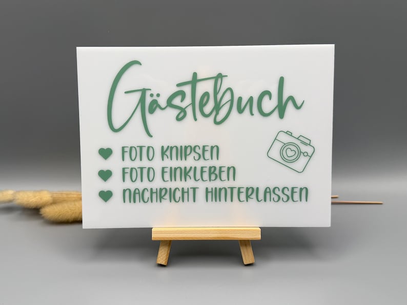 May include: A rectangular white acrylic sign featuring sage green text that reads G&auml;stebuch, Foto knipsen, Foto einkleben, and Nachricht hinterlassen. The sign includes three green heart icons and a camera graphic, displayed on a small wooden easel.